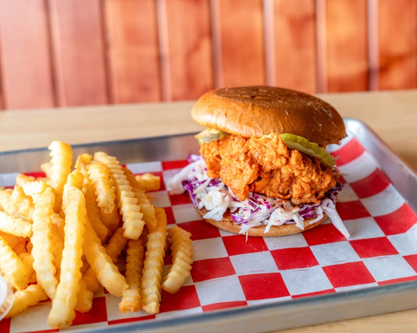 Spicy fried chicken sandwich with coleslaw and pickles, served with crinkle-cut fries on a metal tray.