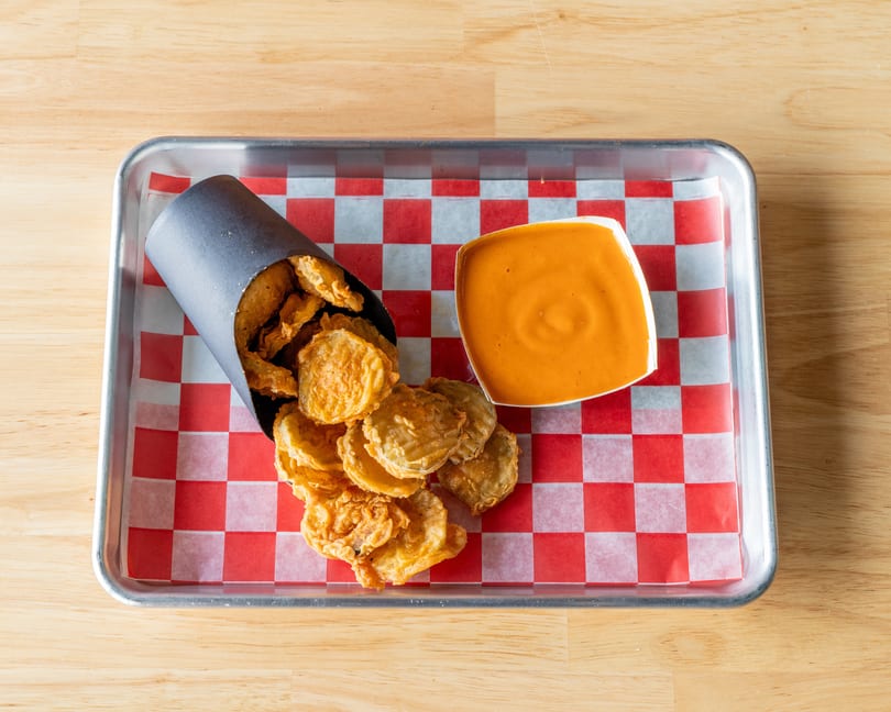 Fried pickles spilling from a black paper cone, with a side of orange dipping sauce on a metal tray.