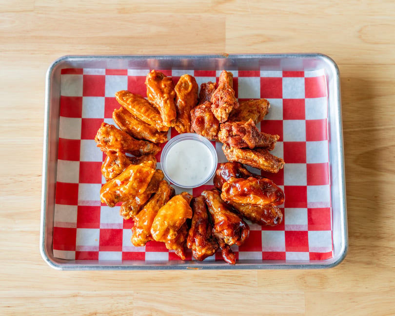 Assortment of chicken wings with various sauces and a side of ranch dressing on a checkered tray.